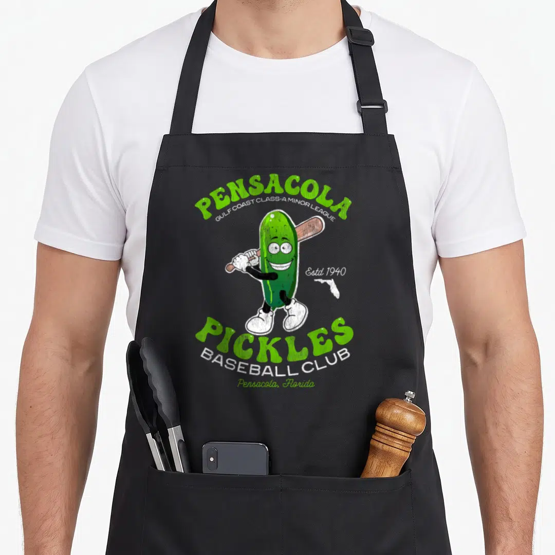 Pensacola pickle apron - Green cartoon pickle holding a brown bat with arching green and white text reading Pensacola — shown on a black cotton canvas apron against a white background