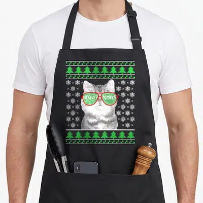 Cat ugly christmas sweater apron - Grey cat in red sunglasses with MEOWY XMAS text and green tree border — shown on a black cotton canvas apron against a white background