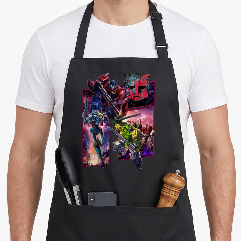 Cybertron transformer apron - Optimus Prime, Bumblebee, and Autobot logo in pink framed panels with red blue yellow — shown on a black cotton canvas apron against a white background