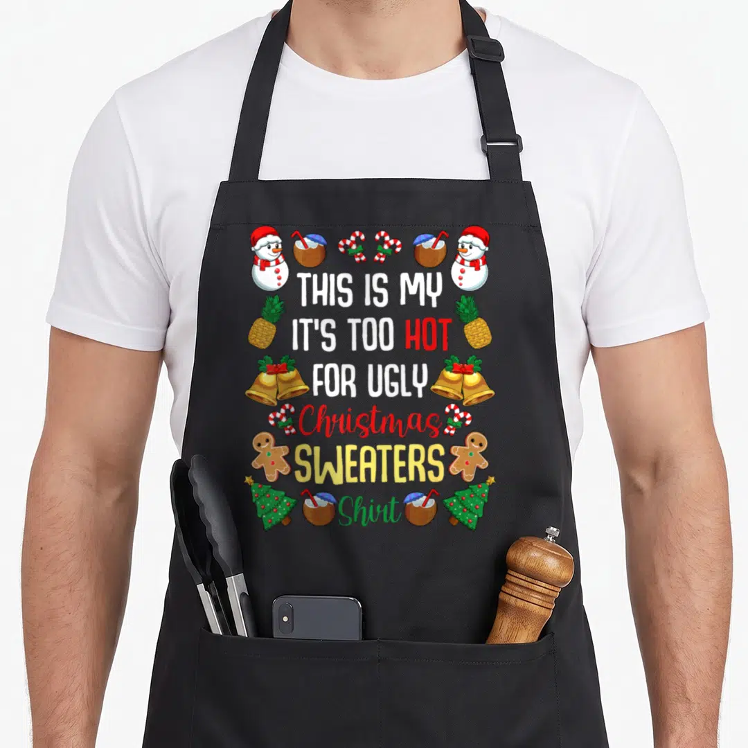 Hot ugly christmas sweater apron - Stacked text surrounded by snowmen, pineapples, gingerbread men, bells, and coconut — shown on a black cotton canvas apron against a white background