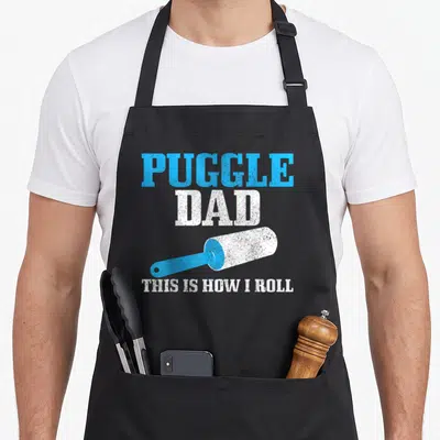 Puggle hair dog apron - Blue and white distressed text PUGGLE DAD THIS IS HOW I ROLL with rolling pin graphic — shown on a black cotton canvas apron against a white background