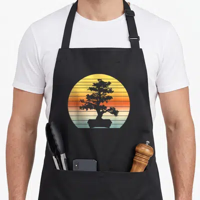 Japanese sun tree apron - Grey bonsai silhouette inside a circle of yellow orange and light blue horizontal stripes — shown on a black cotton canvas apron against a white background