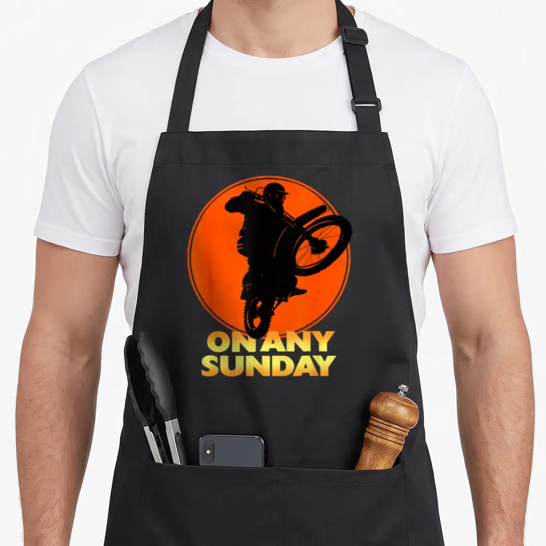 Cute sunday apron - Black motocross rider silhouette on orange circle with yellow ON ANY SUNDAY text — shown on a black cotton canvas apron against a white background