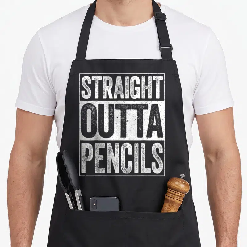Outta pencil apron - Stacked STRAIGHT OUTTA PENCILS text in grey and white blocks inside a rectangular border — shown on a black cotton canvas apron against a white background