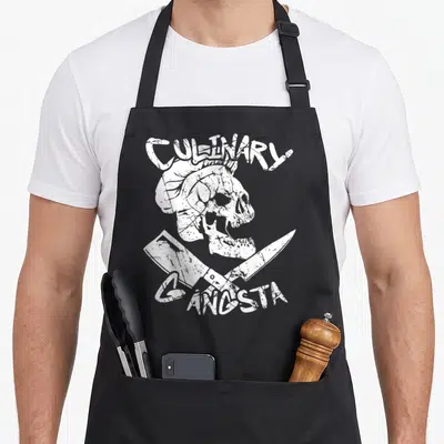 Cute gangster culinary aprons - White distressed skull in chef hat with crossed knives and CULINARY GANGSTA text — shown on a black cotton canvas apron against a white background
