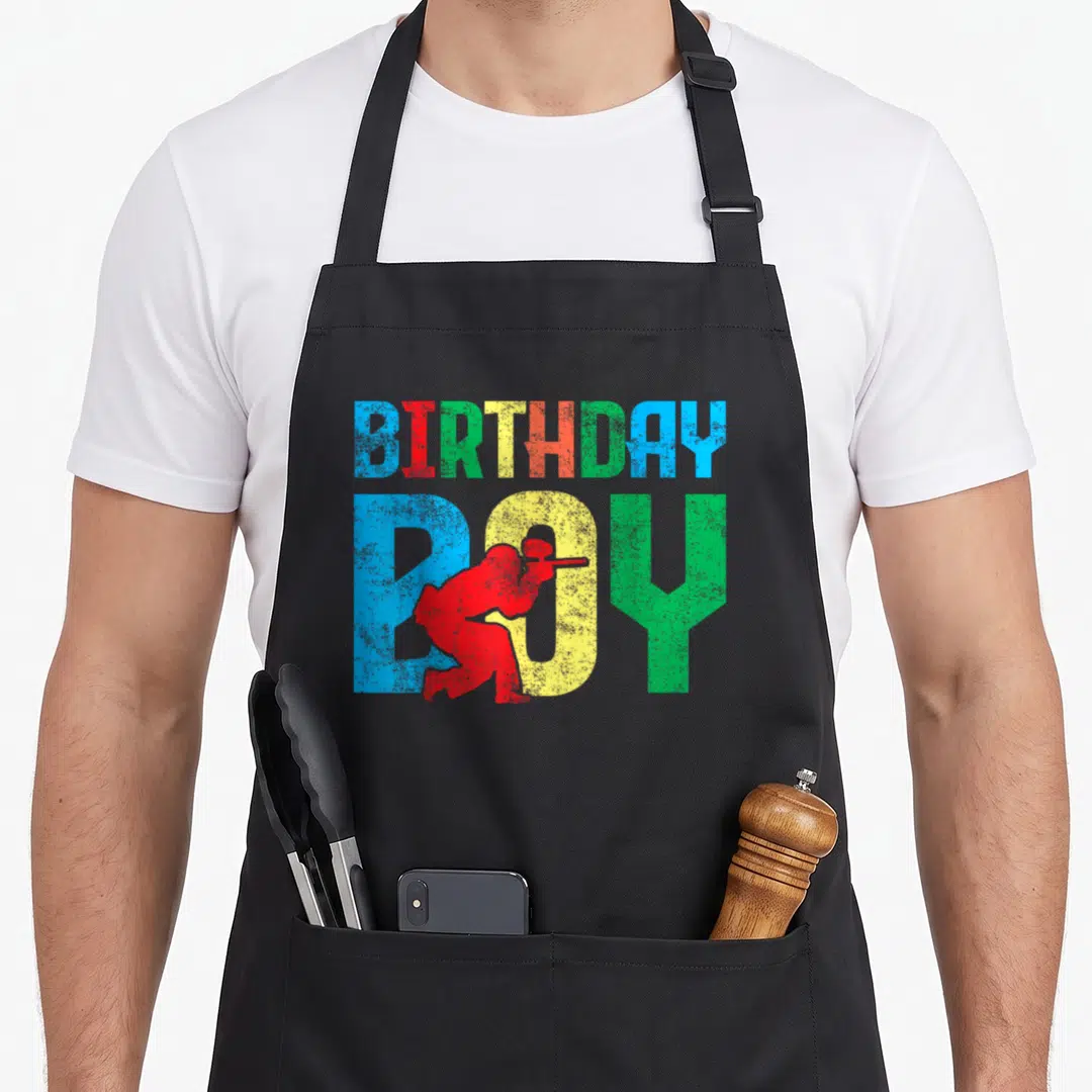 Funny paintball birthday apron - BIRTHDAY BOY text with red hunter silhouette in blue, red, green, and yellow — shown on a black cotton canvas apron against a white background