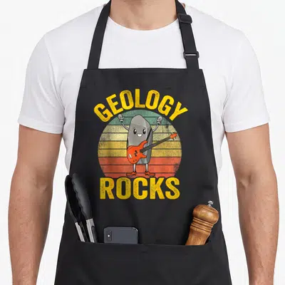 Rock joke apron - Cartoon grey rock playing orange bass guitar with GEOLOGY ROCKS text over retro striped sun — shown on a black cotton canvas apron against a white background