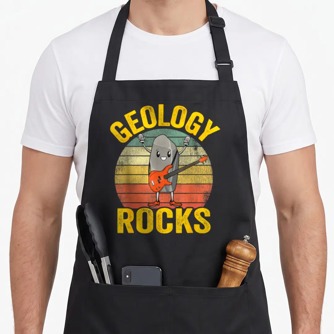 Rock joke apron - Cartoon grey rock playing orange bass guitar with GEOLOGY ROCKS text over retro striped sun — shown on a black cotton canvas apron against a white background