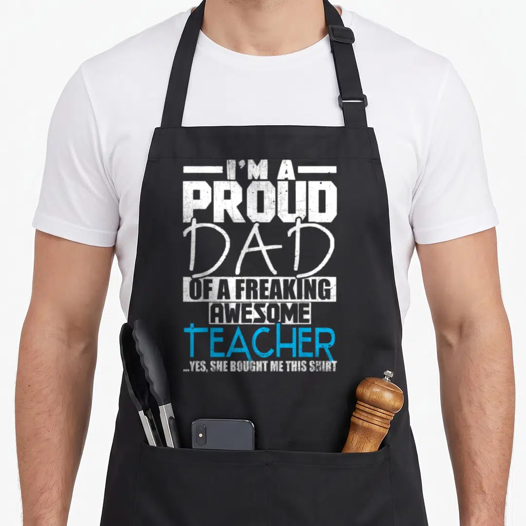 Proud personalized teacher apron - Stacked distressed text reading I'm a proud dad of a freaking awesome teacher — shown on a black cotton canvas apron against a white background