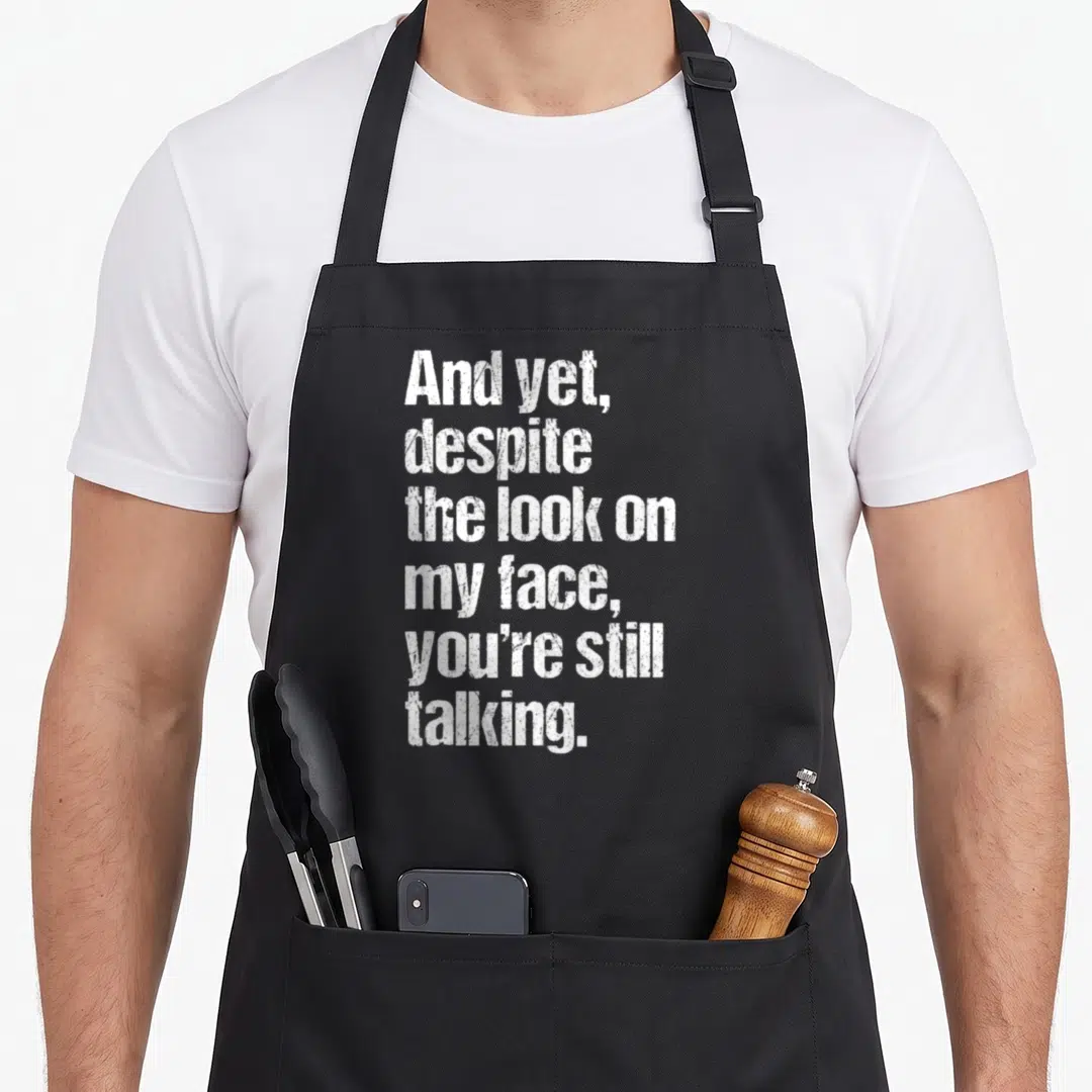 Trending sarcastic apron - White distressed text reading And yet despite the look on my face you're still talking — shown on a black cotton canvas apron against a white background