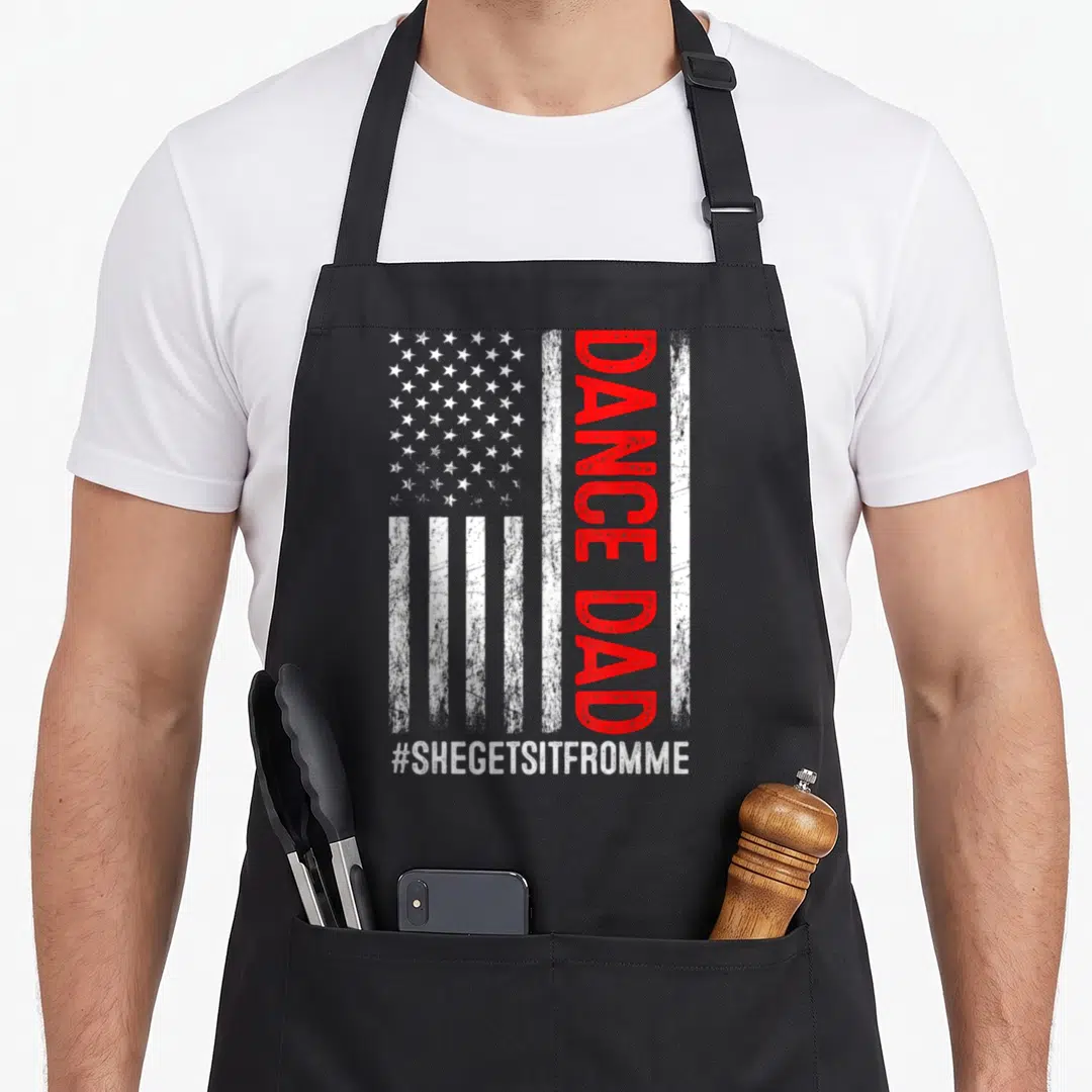 She dance quotes apron - Distressed USA flag with vertical red DANCE DAD text and white #SHEGETSITFROMME hashtag below — shown on a black cotton canvas apron against a white background