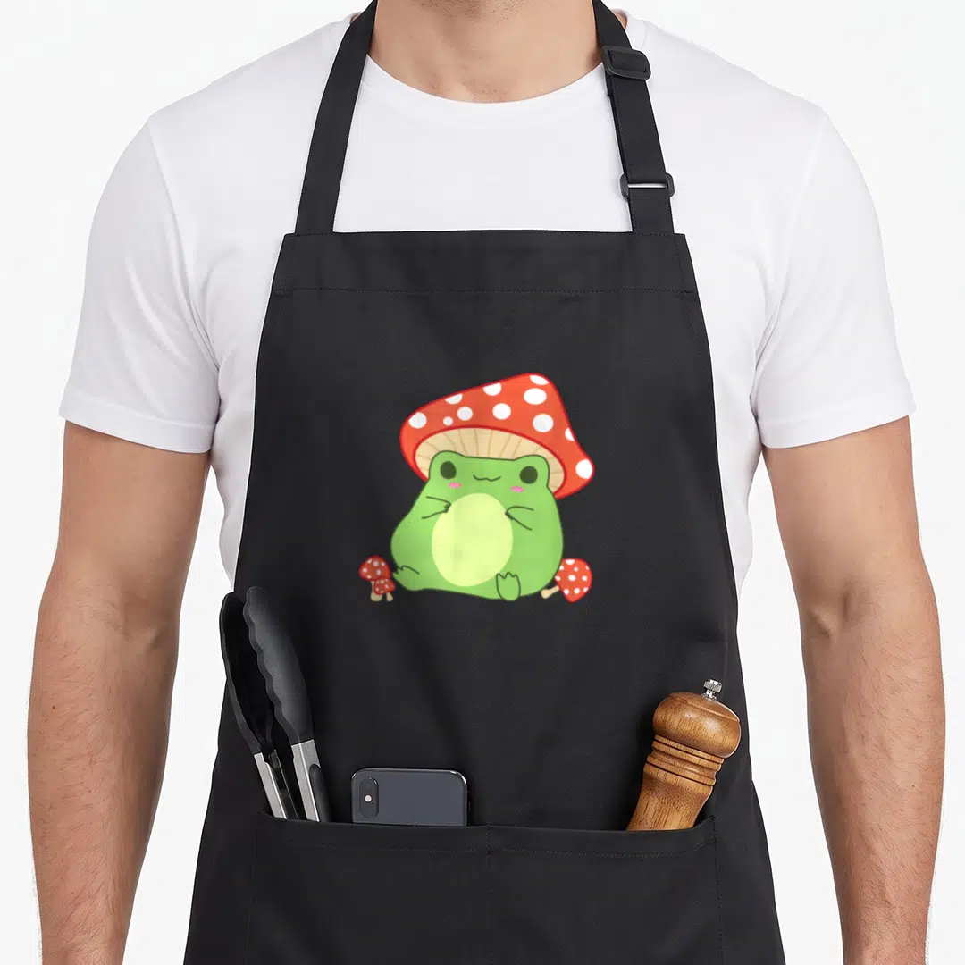 Frog mushroom cottagecore apron - Cartoon green frog wearing red mushroom hat with two smaller mushrooms on ground — shown on a black cotton canvas apron against a white background