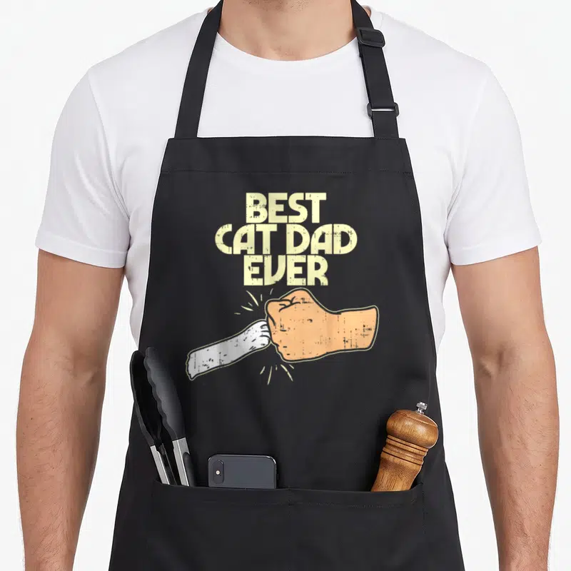 Kitten pet cat apron - Stacked beige text Best Cat Dad Ever above cartoon human and white cat paw fist bump — shown on a black cotton canvas apron against a white background