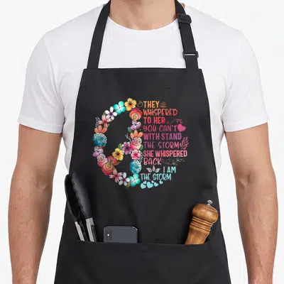 Withstand they whispered apron - C-shaped watercolor floral arrangement in orange, blue, pink, and green framing — shown on a black cotton canvas apron against a white background