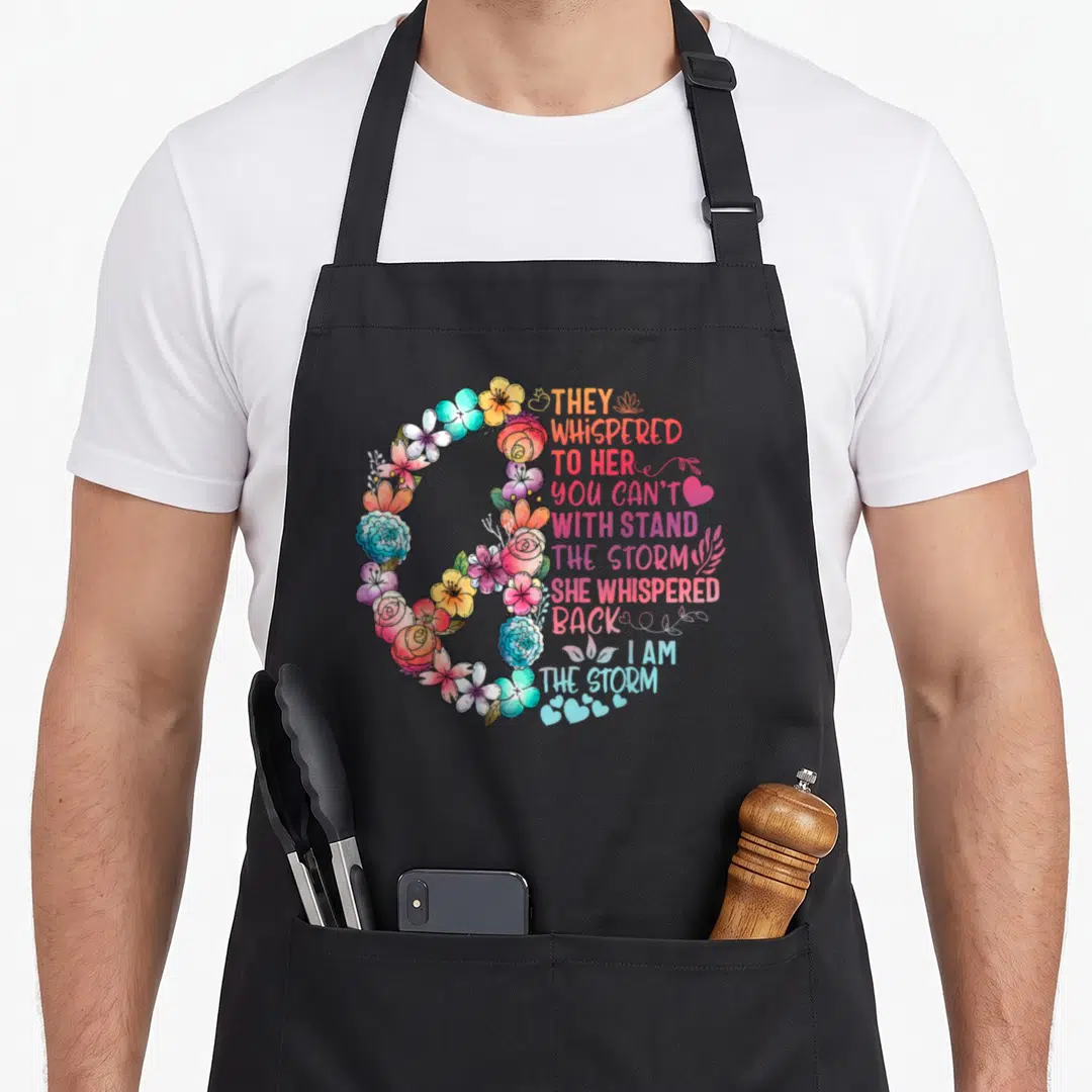 Withstand they whispered apron - C-shaped watercolor floral arrangement in orange, blue, pink, and green framing — shown on a black cotton canvas apron against a white background