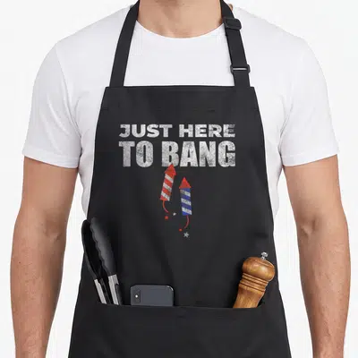 Funny bang 4th of july apron - JUST HERE TO BANG text with two red and blue firework rockets and scattered stars — shown on a black cotton canvas apron against a white background