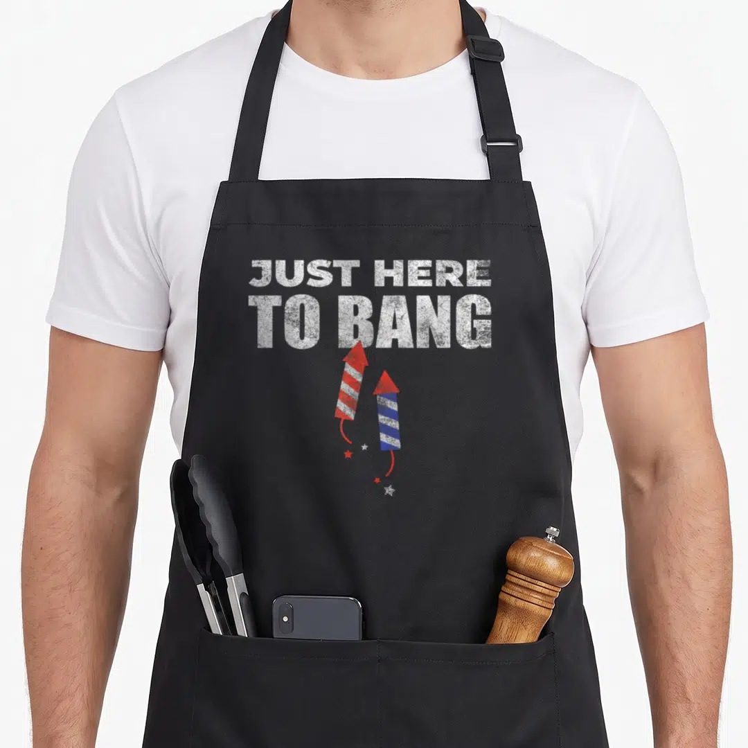 Funny bang 4th of july apron - JUST HERE TO BANG text with two red and blue firework rockets and scattered stars — shown on a black cotton canvas apron against a white background
