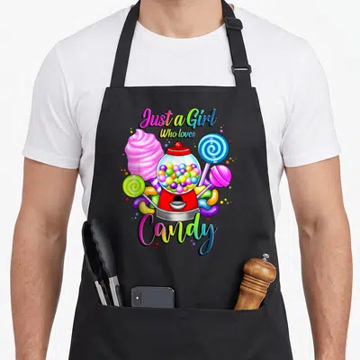 Rainbow cotton candy apron - Cartoon gumball machine with pink cotton candy, lollipops, and rainbow text Just a Girl — shown on a black cotton canvas apron against a white background