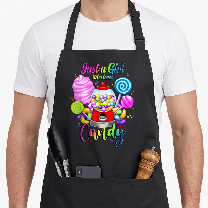 Rainbow cotton candy apron - Cartoon gumball machine with pink cotton candy, lollipops, and rainbow text Just a Girl — shown on a black cotton canvas apron against a white background