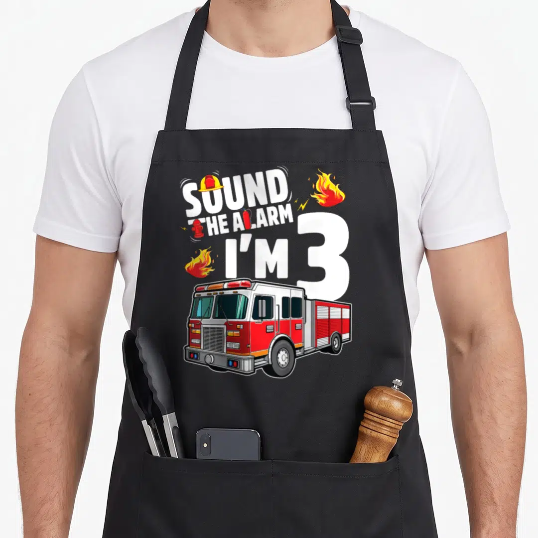 Fire 3rd truck apron - SOUND THE ALARM I'M 3 text with fire truck, flames, helmet, hydrant, and extinguisher in red — shown on a black cotton canvas apron against a white background