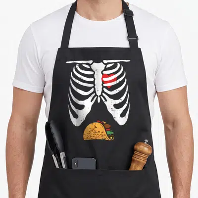 Taco halloween apron - Distressed white ribcage with red heart and yellow taco illustration — shown on a black cotton canvas apron against a white background