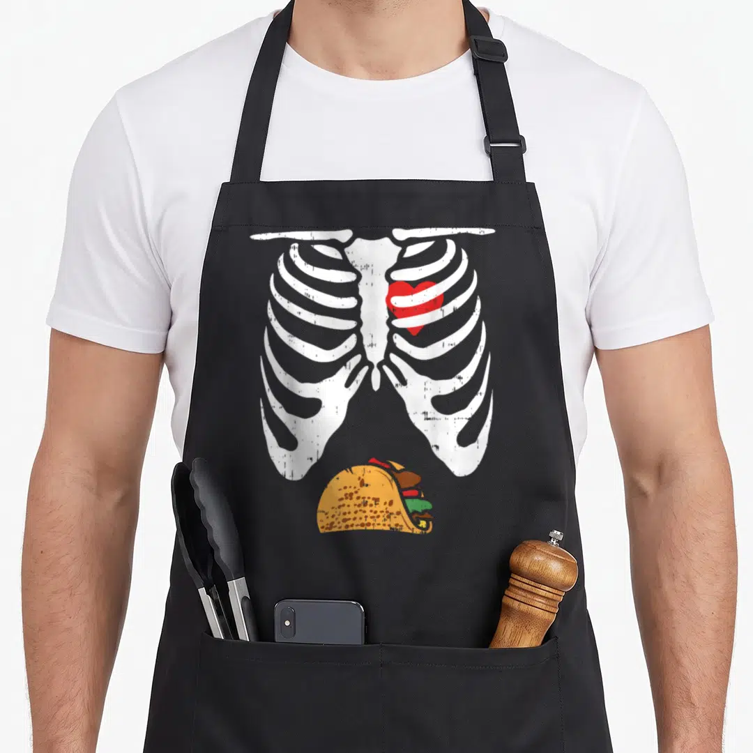 Taco halloween apron - Distressed white ribcage with red heart and yellow taco illustration — shown on a black cotton canvas apron against a white background