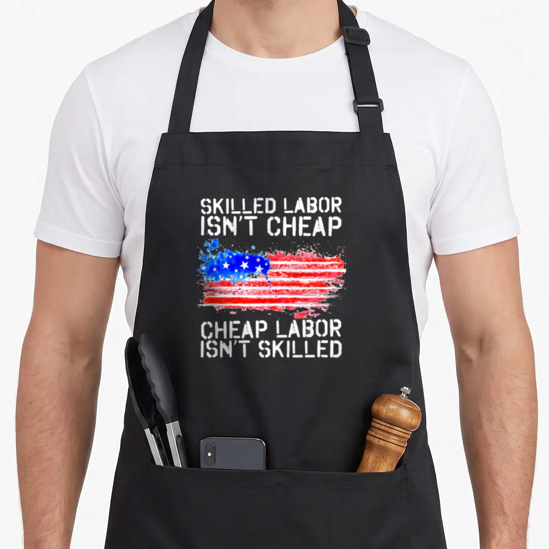 Skilled labor american flag apron - Distressed USA flag with stenciled text Skilled Labor Isn't Cheap Cheap Labor Isn't — shown on a black cotton canvas apron against a white background
