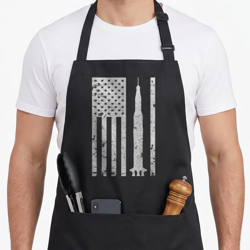 Saturn apollo apron - White distressed rocket and stylized US flag graphic on canvas apron — shown on a black cotton canvas apron against a white background