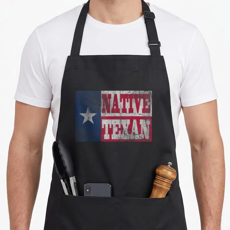 Flag texans apron - Distressed Native Texan text over blue white red Texas flag pattern — shown on a black cotton canvas apron against a white background