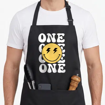 1st custom face apron - Yellow smiley face with lightning eyes over stacked white ONE text — shown on a black cotton canvas apron against a white background