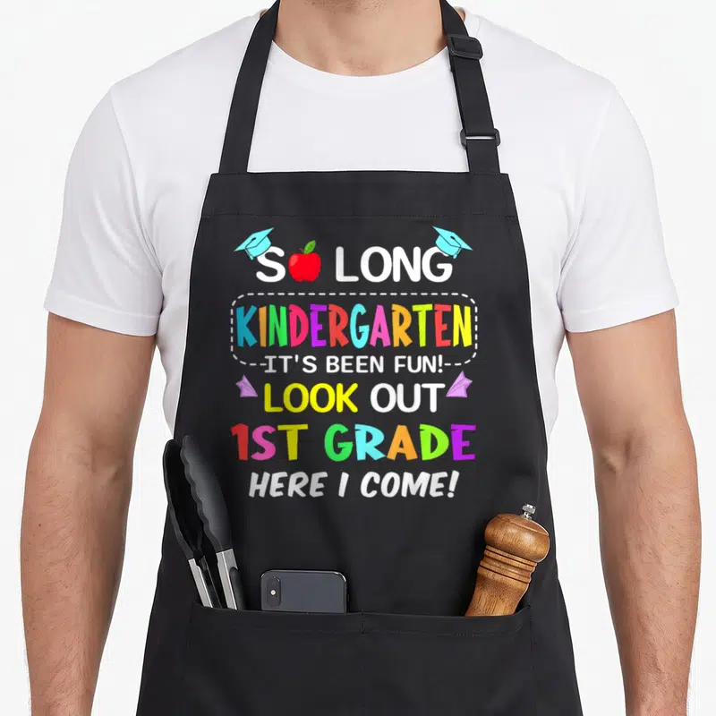 1st come and get it apron - Stacked colorful text reading So Long Kindergarten 1st Grade Here I Come with apple — shown on a black cotton canvas apron against a white background