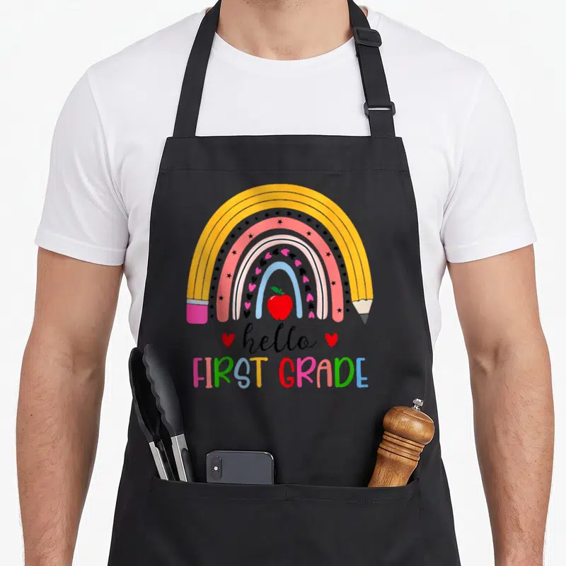 Hello grade teacher apron - Rainbow pencil design with stars hearts and apple text reading hello FIRST GRADE — shown on a black cotton canvas apron against a white background