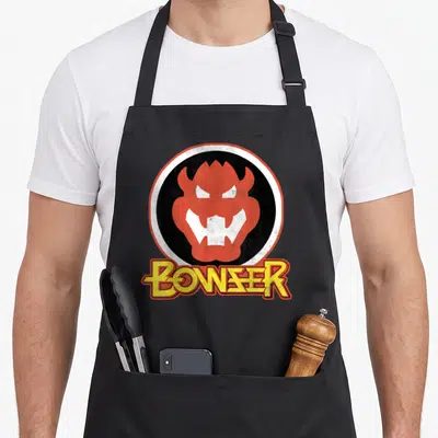 Portrait holiday apron - Orange Bowser head in black circle with white border, yellow BOWSER text below — shown on a black cotton canvas apron against a white background