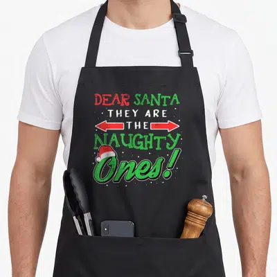 Dear christmas santa apron - Stacked text DEAR SANTA THEY ARE THE NAUGHTY Ones with Santa hat and double arrow in red — shown on a black cotton canvas apron against a white background