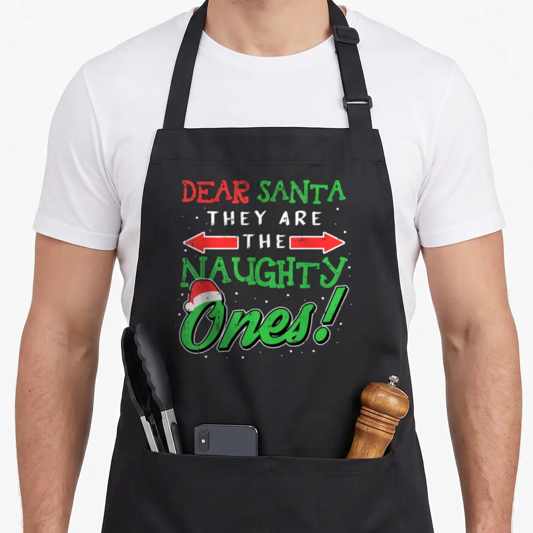 Dear christmas santa apron - Stacked text DEAR SANTA THEY ARE THE NAUGHTY Ones with Santa hat and double arrow in red — shown on a black cotton canvas apron against a white background