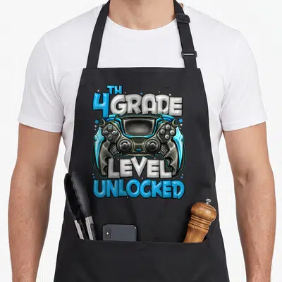 4th game aprons - Game controller with TH 4 GRADE LEVEL UNLOCKED text and scattered stars in blue and white — shown on a black cotton canvas apron against a white background