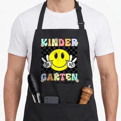 Smile face apron - Yellow smiley face with two peace sign hands over black white checkerboard, colorful KINDER GARTEN — shown on a black cotton canvas apron against a white background