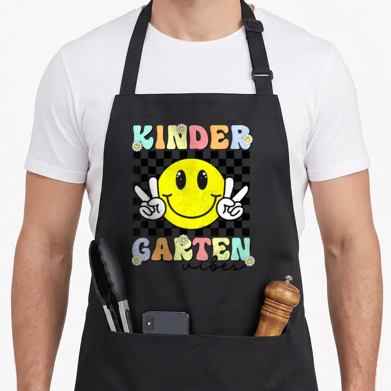 Smile face apron - Yellow smiley face with two peace sign hands over black white checkerboard, colorful KINDER GARTEN — shown on a black cotton canvas apron against a white background