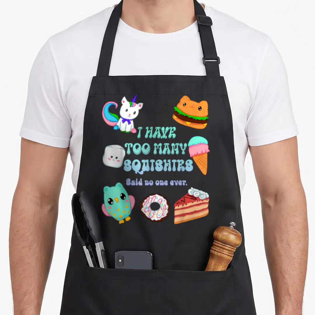 Many grade dumpling apron - I HAVE TOO MANY SQUISHIES text surrounded by cartoon unicorn, cat burger, and ice cream — shown on a black cotton canvas apron against a white background