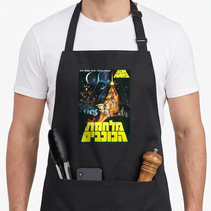 Movie hebrew aprons - Vintage Star Wars poster with Hebrew text, Luke Skywalker, and space ships in yellow and blue — shown on a black cotton canvas apron against a white background
