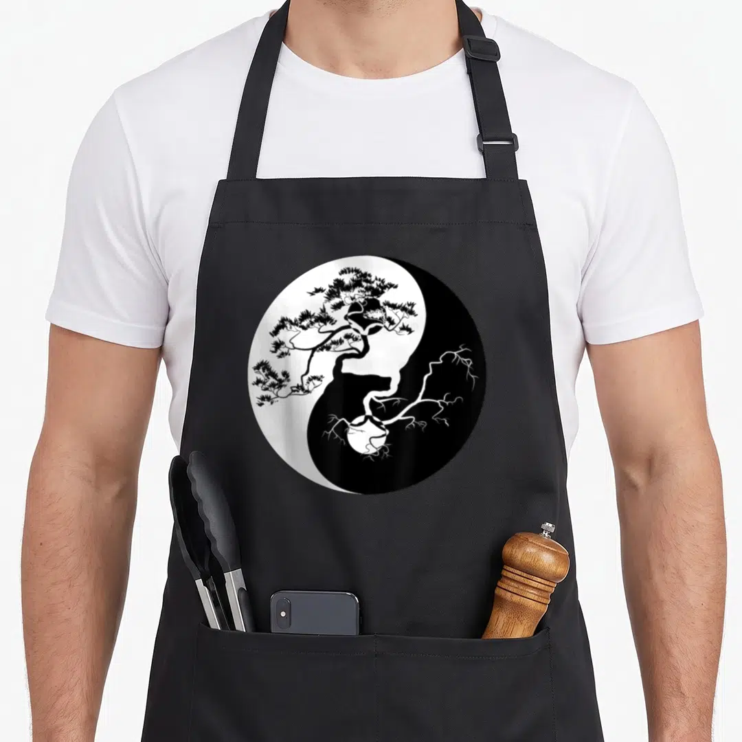 Yin tree apron - Black and white yin yang symbol containing a pine tree with foliage above and roots below — shown on a black cotton canvas apron against a white background