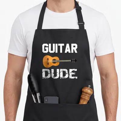 Classical acoustics apron - GUITAR DUDE text flanking an orange cartoon guitar in white and black ink — shown on a black cotton canvas apron against a white background