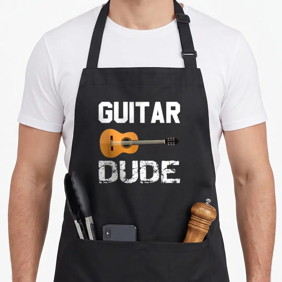 Classical acoustics apron - GUITAR DUDE text flanking an orange cartoon guitar in white and black ink — shown on a black cotton canvas apron against a white background
