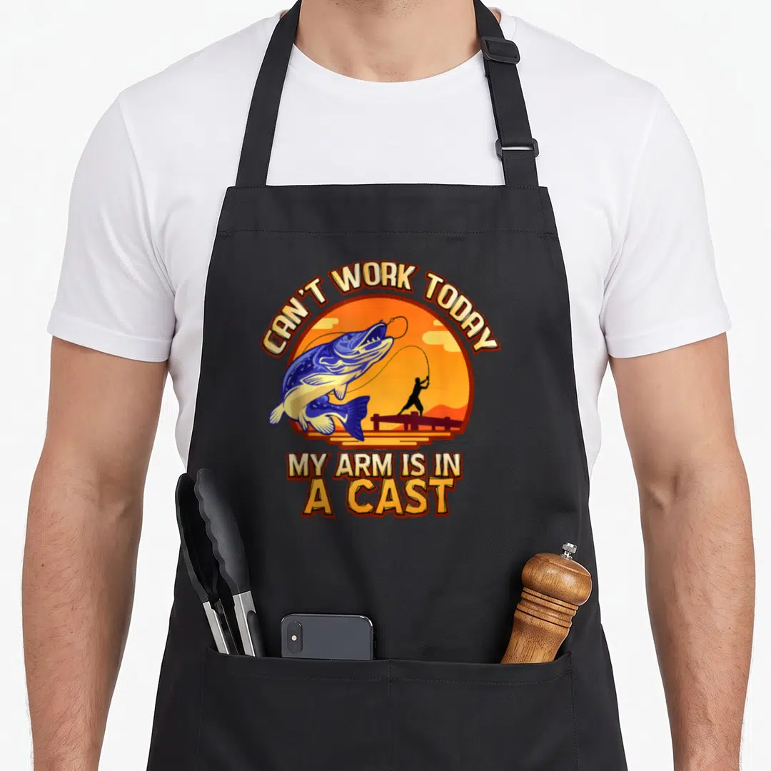 Reeling fishing aprons - Can't Work Today My Arm Is In A Cast jumping fish pun design — shown on a black cotton canvas apron against a white background
