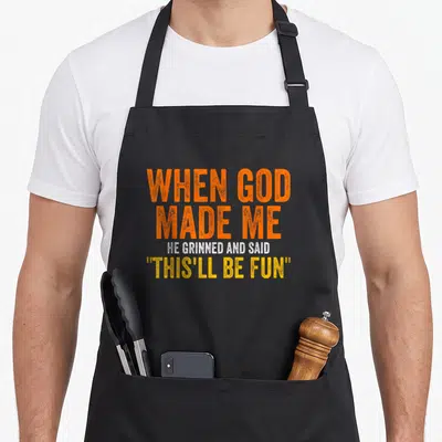 Funny god funny christian apron - Stacked orange and yellow typography reading When God Made Me He Grinned And Said — shown on a black cotton canvas apron against a white background