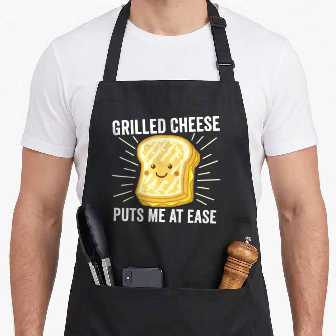 Grilled food aprons - Cartoon grilled cheese sandwich with smiling face and radiating lines under GRILLED CHEESE PUTS — shown on a black cotton canvas apron against a white background
