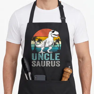 Unclesaurus t rex apron - T-Rex in sunglasses over retro striped sun with Uncle Saurus text — shown on a black cotton canvas apron against a white background