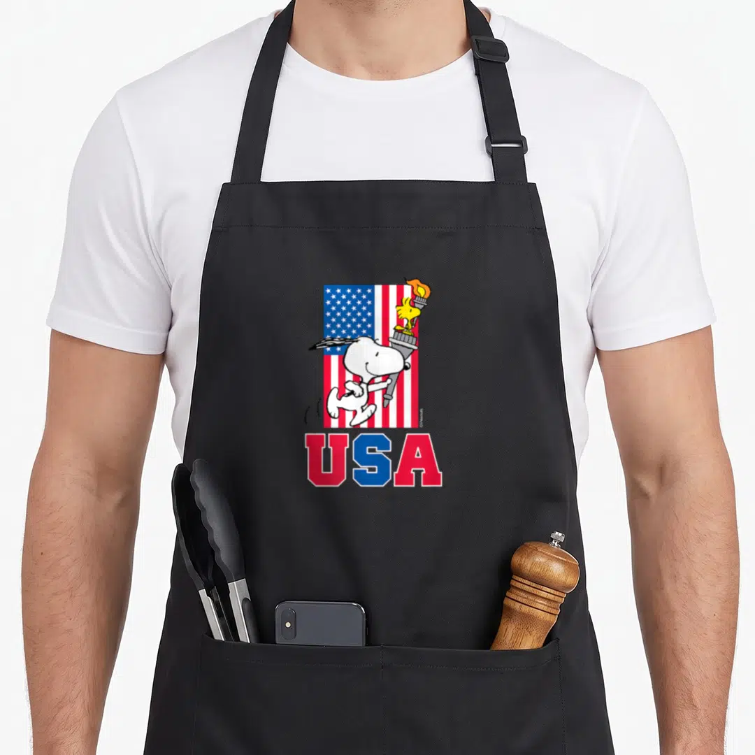 Woodstock flag apron - Snoopy running with torch and Woodstock over red white blue USA flag cartoon art — shown on a black cotton canvas apron against a white background
