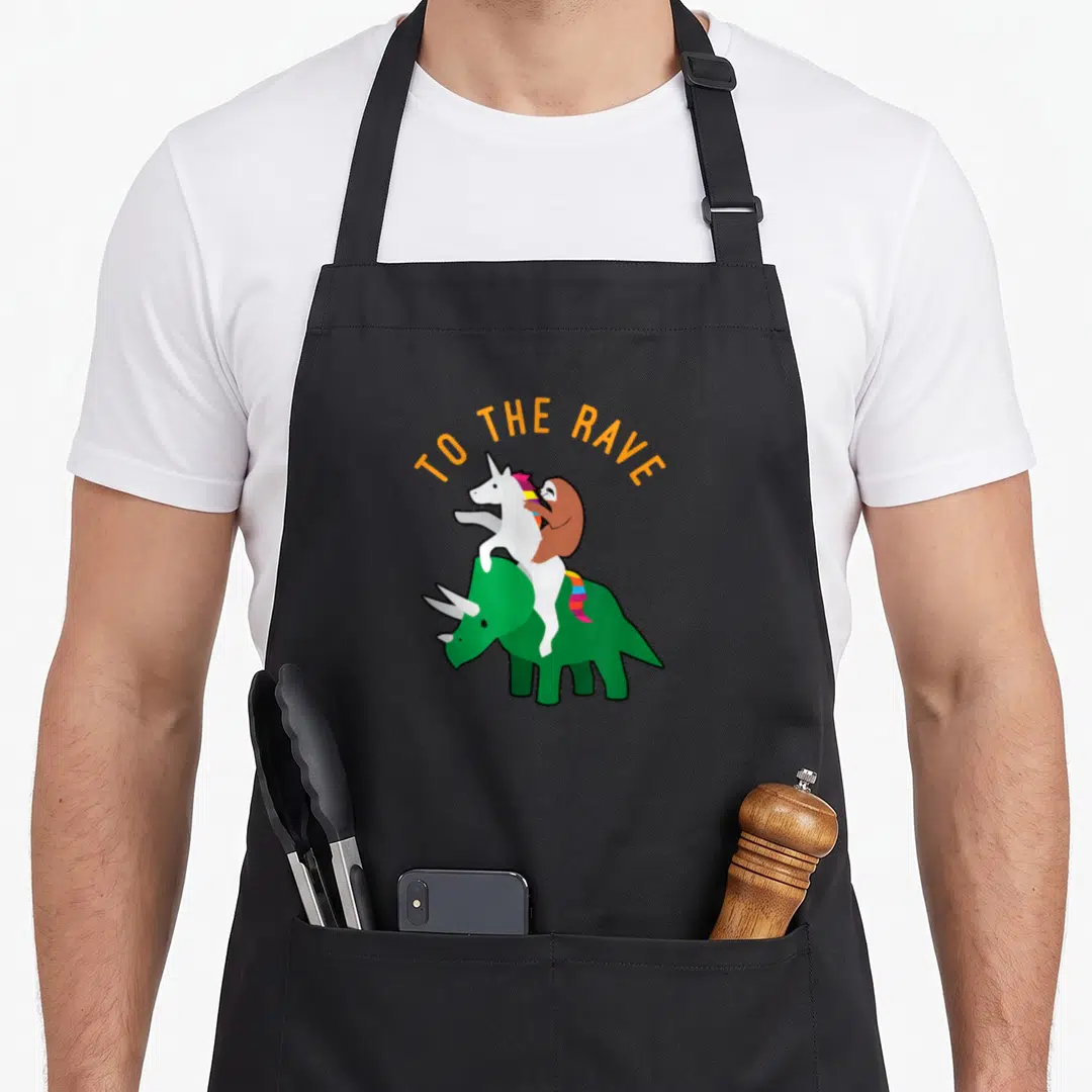Rave dinosaur apron - Orange TO THE RAVE text over green triceratops carrying white unicorn and brown sloth — shown on a black cotton canvas apron against a white background