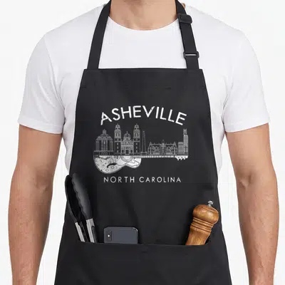 Souvenir asheville apron - White distressed Asheville skyline integrated into a guitar body shape with North Carolina — shown on a black cotton canvas apron against a white background